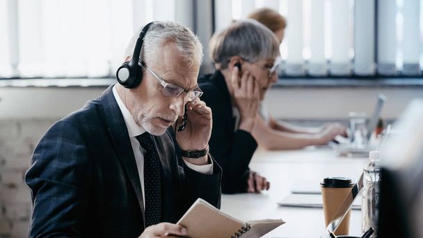 Mature businessman in headset looking at notebook near laptop in office  - Photo, Image