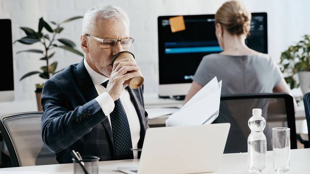 Mature businessman drinking coffee to go and holding papers near laptop in office  - Фото, зображення