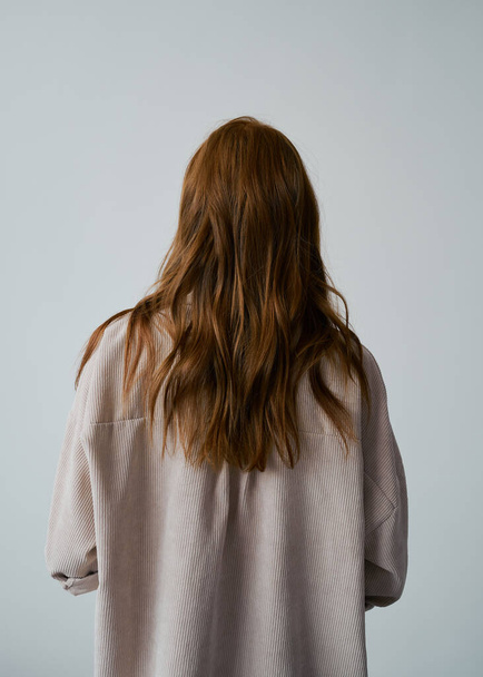 Portrait of young amazing redhead girl in beige shirt on neutral background. Long wavy healthy hair. Beautiful and fresh. Natural beauty. Minimalism. Soft light. Good quality. Hair advertising. No face. - Photo, Image