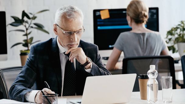Empresário em óculos escrevendo no caderno e olhando para laptop perto colega borrado no escritório  - Foto, Imagem