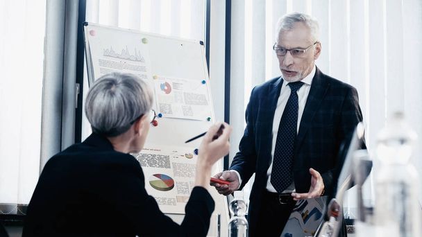 Businessman in formal wear talking to blurred colleague near flip chart in office  - Photo, image