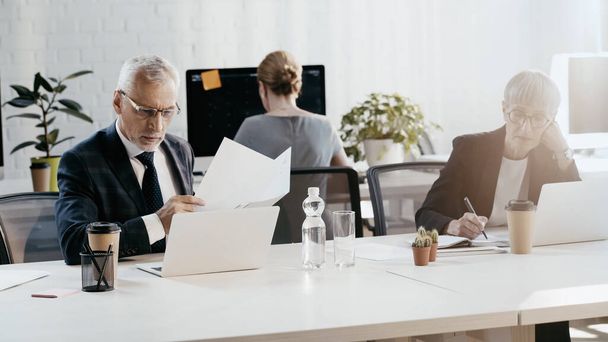 Mature businessman holding documents near laptop and colleague working in office  - Photo, Image