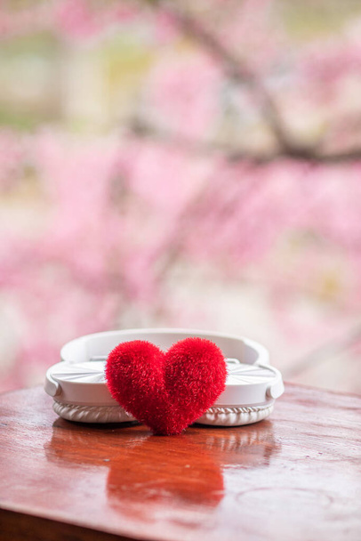 rotes Herz in der Hand auf verschwommenem Hintergrund aus rosafarbenen Kirschblüten sieht schön aus und rote Herzen sind für Verliebte am Valentinstag vorbereitet, da es ausdrückt, was sie einander schenken wollen. - Foto, Bild
