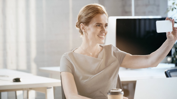 Positive businesswoman taking selfie on smartphone near laptop in office  - Valokuva, kuva