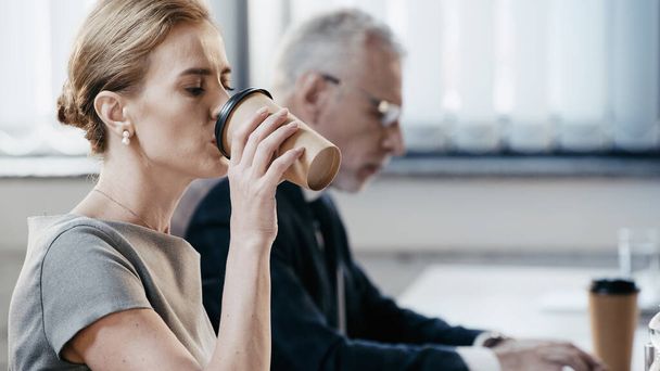 Businesswoman drinking coffee to go near blurred colleague working in office  - Foto, imagen
