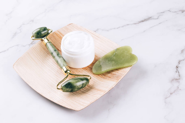 Jade facial gua sha roller with moisturizing face cream on white marble table background with copy space. Facial massage kit for lifting massage therapy. Mock up. Selective focus - Zdjęcie, obraz