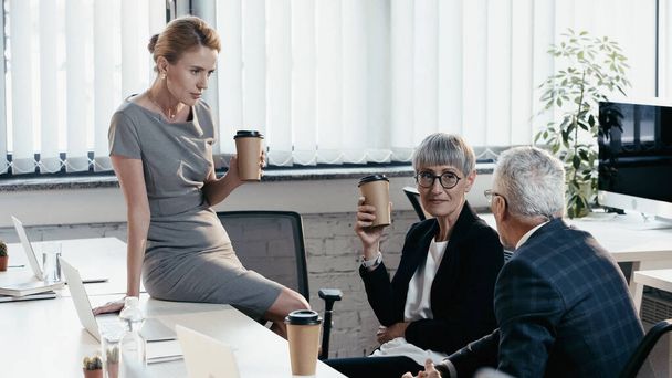 Менеджеры в формальной одежде держат чашки на вынос рядом с ноутбуками в офисе  - Фото, изображение