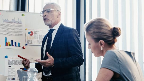Mature businessman talking near flip chart and blurred businesswoman in office  - Photo, Image