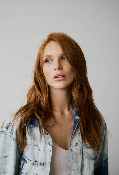 Nice model with red hair, blue eyes and long wavy red hair. Sensual attractive woman. Freckles. Neutral background. Excellent quality. Soft light. Jeans jacket. Nice lashes, big lips, beautiful eyes - Foto, Imagem