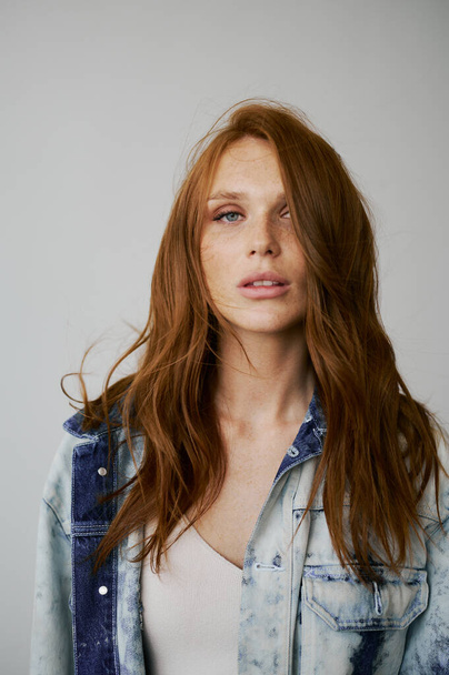 Nice model with red hair, blue eyes and long wavy red hair. Sensual attractive woman. Freckles. Neutral background. Excellent quality. Soft light. Jeans jacket. Nice lashes, big lips, beautiful eyes - Фото, изображение