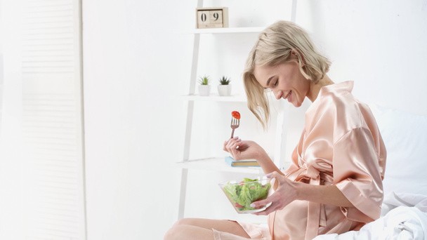 cheerful pregnant woman holding bowl with salad and looking at belly in bedroom  - Foto, immagini
