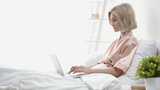 freelance biondo usando il computer portatile mentre si siede a letto - Foto, immagini
