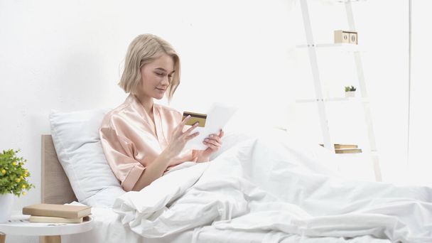 femme souriante tenant tablette numérique et carte de crédit tout en faisant des achats en ligne au lit  - Photo, image