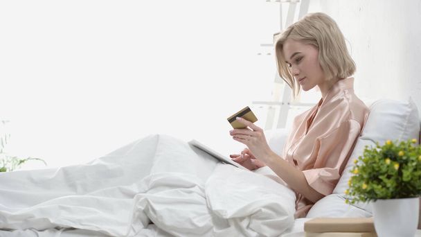ベッドでデジタルタブレットとクレジットカードを持っているブロンドの女性の側の見解  - 写真・画像