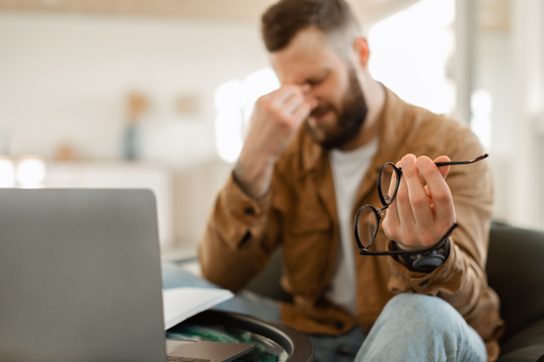 Tired Male Holding Glasses Massaging Nosebridge Sitting At Laptop Indoors - Foto, immagini