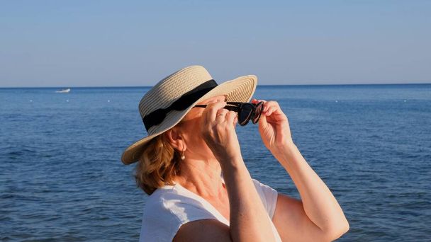 青い海の背景にわらの帽子とサングラスを着用し、成熟した女性、年齢50の肖像画。夏休み休暇現役引退者 - 写真・画像