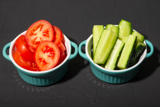 Sliced cucumbers and tomatoes in cups on a dark background. Salad ingredients. Minimalism. Raw vegetables, vegetarianism. - Photo, Image
