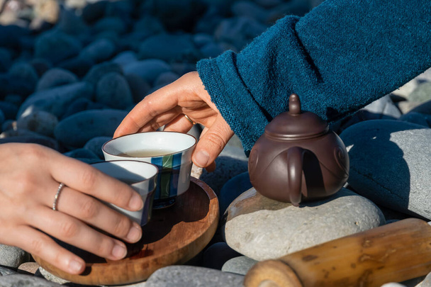 Крупный план рук двух женщин взять чашки чая из деревянного подноса, есть специальный чайник рядом с ним. Чайная церемония на галечном пляже моря в солнечный день - Фото, изображение
