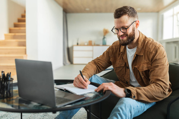 Επιτυχημένος επιχειρηματίας κρατώντας σημειώσεις στο φορητό υπολογιστή που κάθεται στο σπίτι - Φωτογραφία, εικόνα