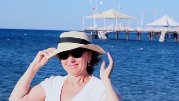 Olgun bir kadının portresi, 50 yaşında, hasır şapka ve güneş gözlüğü takıyor mavi bir deniz arka planında. Yaz, tatil, tatil, aktif emekliler - Video, Çekim
