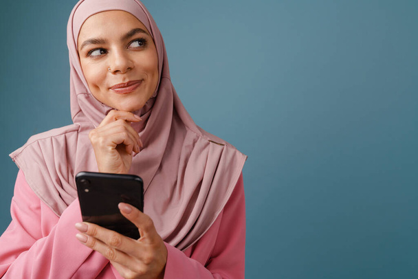 Jeune femme musulmane en hijab souriant en utilisant un téléphone portable isolé sur fond bleu - Photo, image