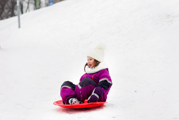 Linda niña divertida en un mono púrpura y un sombrero con un pompón en el parque paseos en una pista de hielo rojo brillante durante una nevada. Actividades al aire libre de invierno - Foto, Imagen