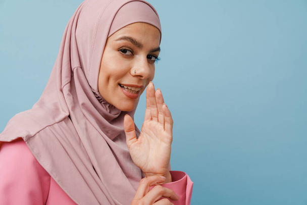 Young muslim woman in hijab gesturing while whispering isolated over blue background - Фото, изображение