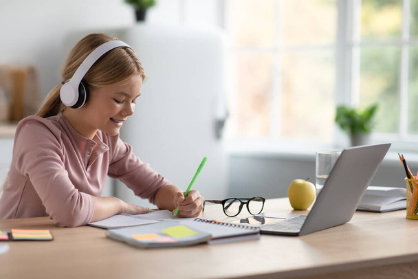 Ευτυχισμένος καυκάσιος έφηβος κορίτσι ξανθιά στα ακουστικά κρατά σημειώσεις, μελέτη στο σπίτι στο τραπέζι με φορητό υπολογιστή στην κουζίνα - Φωτογραφία, εικόνα
