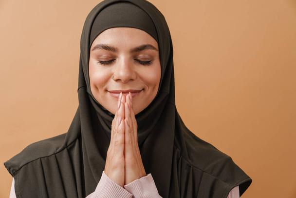 Young muslim woman in hijab smiling and making pray gesture isolated over beige background - Фото, изображение