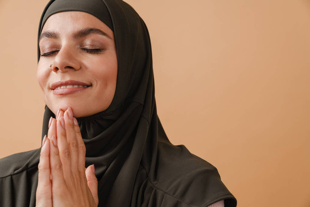 Молодая мусульманка в хиджабе улыбается и делает молитвенный жест изолированным на бежевом фоне - Фото, изображение