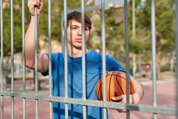 Genç bir sporcu elinde basketbol topuyla basketbol sahasında metal bir çite yaslanıyor. - Fotoğraf, Görsel