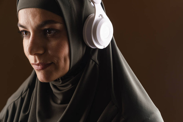 Молодая мусульманка в хиджабе слушает музыку с наушниками на коричневом фоне - Фото, изображение