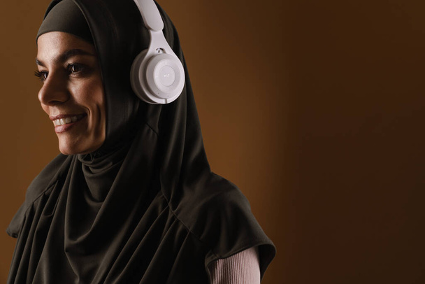 Young muslim woman in hijab listening music with headphones isolated over brown background - Photo, image