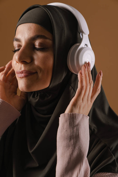 Young muslim woman in hijab listening music with headphones isolated over brown background - Foto, imagen