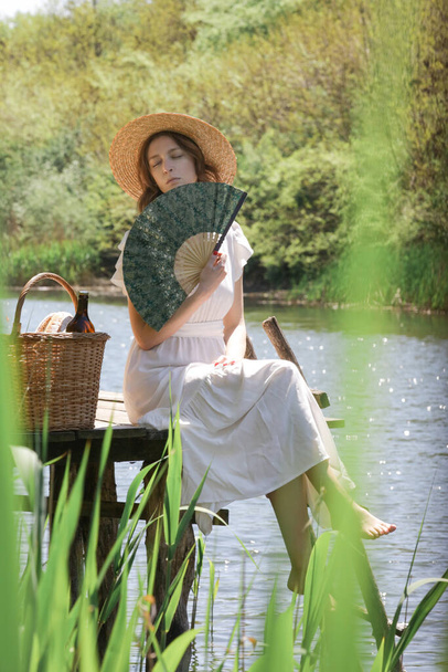 Νεαρή γυναίκα με μακρύ λευκό φόρεμα και ψάθινο καπέλο κάθεται στην προβλήτα και κοιτάζει το ποτάμι. Ευαίσθητες στιγμές στη φύση, αργή και απλή διαβίωση. - Φωτογραφία, εικόνα