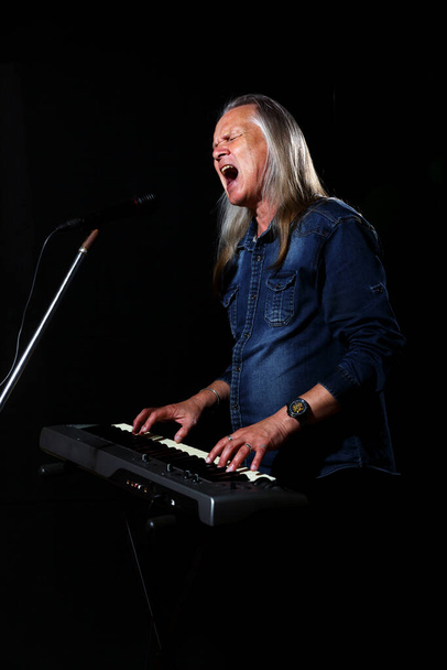 homem idoso com cabelos grisalhos longos em camisa jeans azul joga as chaves e canta com microfone no palco em fundo preto - Foto, Imagem