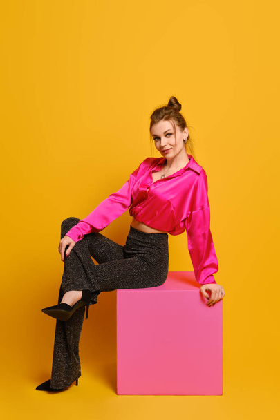Playful woman in bright old-fashioned clothes sits in profile on purple cube over bright yellow studio background - Foto, Imagem