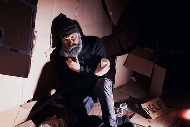 Un hombre sin hogar barbudo se sienta en cajas en la calle y pide ayuda. ¿Necesita una persona sin hogar pide dinero para la comida y durante la noche. - Foto, imagen