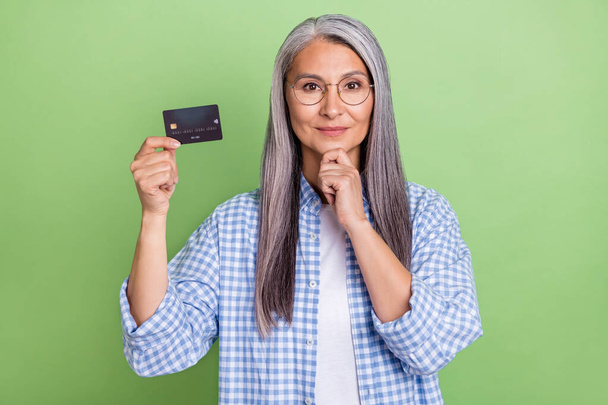 Photo of clever aged white hairdo lady hold card wear eyewear plaid shirt isolated on green color background - 写真・画像