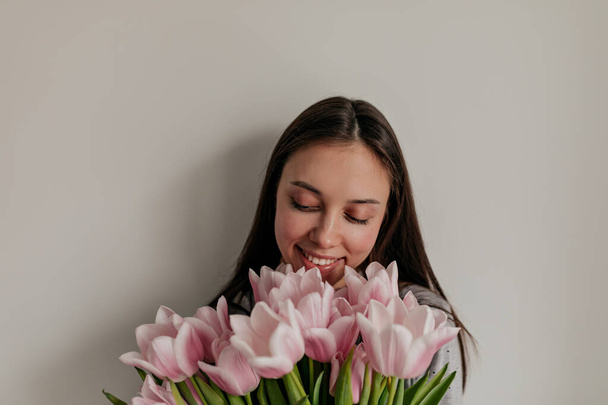 Close up portrait of charming pretty girl with nude make up and dark long hair smiling with close eyes and holding big pink flowers. Happy girl in Woman's Day . High quality photo - Photo, Image