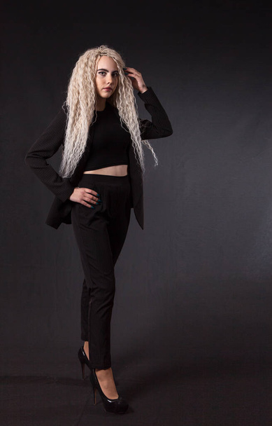 jong blond meisje met lange krullend haar in zwart pak poseren in de studio met zwarte achtergrond - Foto, afbeelding