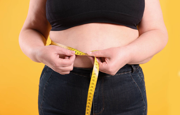 Overweight woman measuring waist with tape on yellow background, closeup - Valokuva, kuva