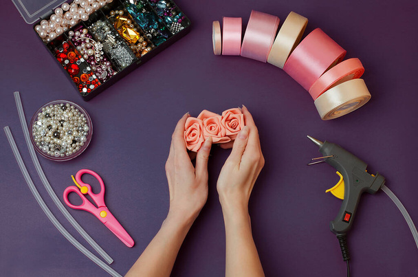 In women's hands,a hairpin with their own hands.The craft tools are set against a dark purple background. Satin pink ribbons, rhinestones, beads, scissors, glue gun, threads, glue sticks.DIY. Handmade - Foto, Imagem