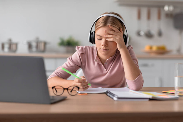 Κουρασμένος απασχολημένος ευρωπαϊκή νέοι κορίτσι ξανθιά με ακουστικά που σπουδάζουν στο σπίτι στο τραπέζι με φορητό υπολογιστή - Φωτογραφία, εικόνα