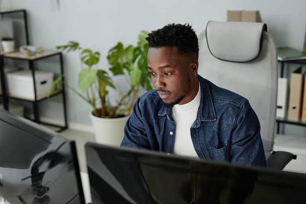 Młody czarny mężczyzna w luźnych ubraniach siedzi przed monitorami komputerowymi - Zdjęcie, obraz