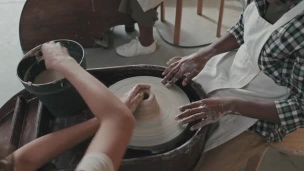 Высокий угол обрезанного черного мальчика, сидящего за гончарным кругом и учащегося делать глиняные изделия в мастерской днем, неузнаваемого учителя, помогающего студенту - Кадры, видео