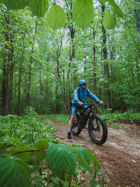 An adult man on an electric bike with thick wheels. The concept of outdoor sports in any weather. Fat bike in the forest in the rain. - Photo, Image