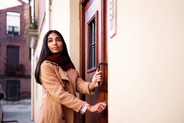 jovem mulher indiana feliz abrindo sua porta de casa e olhando para trás com alegria, espaço cópia - Foto, Imagem