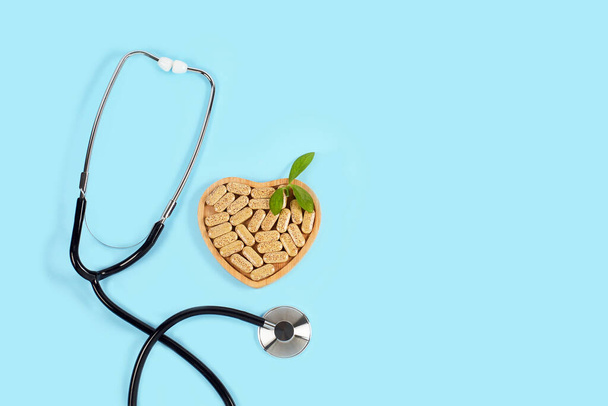 Herbal pills in heart shaped wooden plate and stethoscope on blue background. - Photo, Image