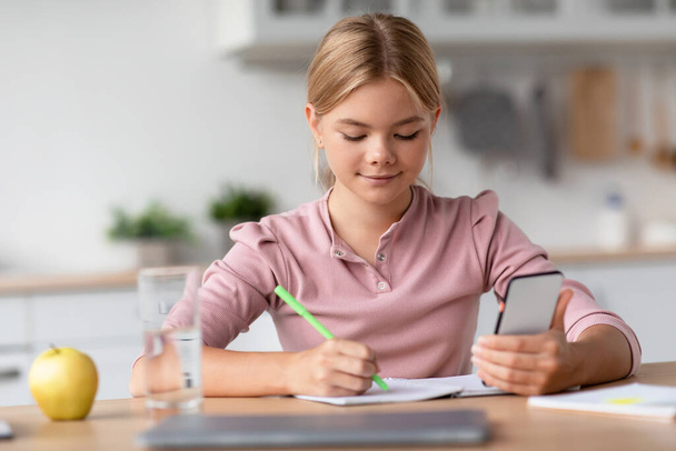 Ernstig mooi blond europees tiener meisje maakt notities met smartphone aan tafel in minimalistische keuken - Foto, afbeelding